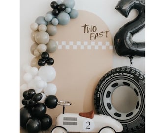 May include: A white toy car with a black steering wheel and a brown leather seat. The car has a black number "2" on the side. The car is in front of a brown and white checkered background with the words "Two Fast" written on it. There are black, white, and blue balloons in the background.