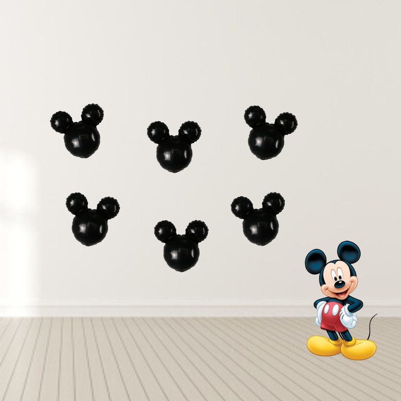 May include: Six black Mickey Mouse head shaped balloons on a white wall with a red and yellow Mickey Mouse character standing on a wood floor.
