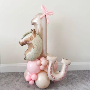 Op de afbeelding: Een feestelijke ballonnenopstelling voor een eerste verjaardag. Het bevat een grote ballon met het cijfer "1", een paardenkopballon, een hoefijzerballon en een cluster van roze en crèmekleurige ballonnen. Een roze strik siert de één.