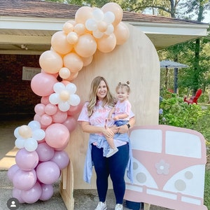 May include: A woman and a young girl stand in front of a wooden backdrop decorated with a balloon arch and a pink and white cutout of a vintage van. The balloon arch is made of pink, white, and peach balloons. The woman is wearing a tie-dye shirt and blue jeans. The girl is wearing a pink and white shirt and blue jeans. The van has a white flower on the side.