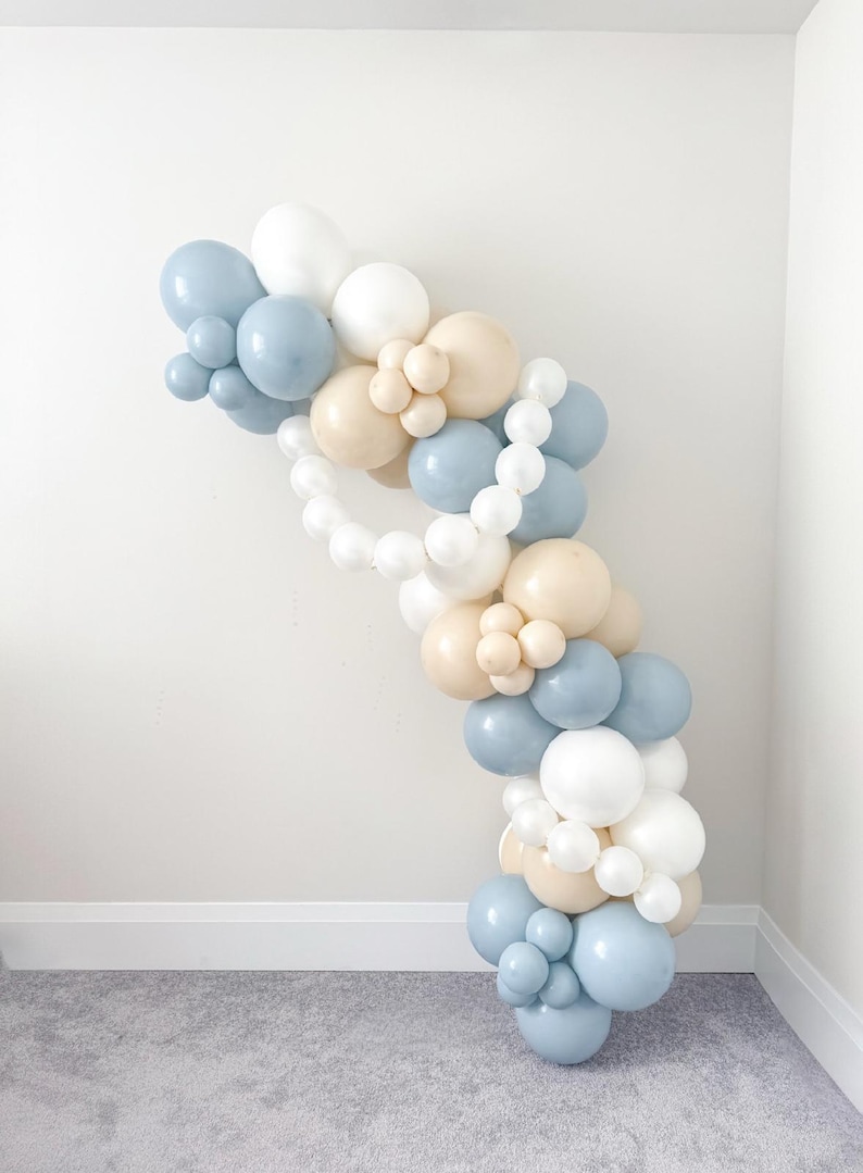 May include: A balloon arch in a soft color palette of white, light blue, and beige. The balloons are arranged in a cascading, curved shape, perfect for a party or event decoration. The balloons are of varying sizes.