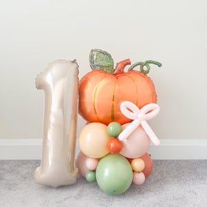 May include: A celebratory balloon display with an orange pumpkin balloon, green stem, and a light pink bow. A champagne-coloured number one balloon is positioned next to the pumpkin. The arrangement includes pastel-coloured balloons.