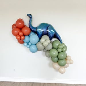 May include: A blue dinosaur balloon with a green, blue, and beige balloon garland. The garland is made of various sizes and shapes of balloons.