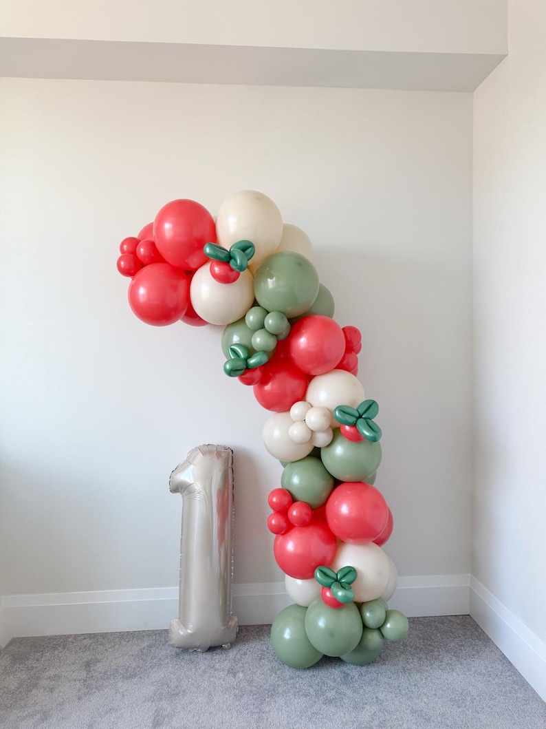 May include: A festive balloon arch in red, white, and green, with decorative accents. A large silver number one balloon stands nearby. The balloon arrangement is likely for a birthday or celebration.