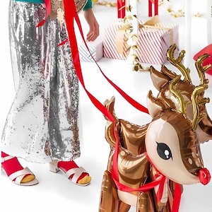 May include: A child in a silver sequined skirt and a turquoise top walks a pair of golden reindeer balloon animals with red ribbons.