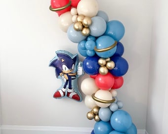 May include: A balloon garland in red, white, blue, and gold, with a Sonic the Hedgehog foil balloon. The garland features various sizes of balloons, some with gold rings, creating a festive and celebratory atmosphere. Perfect for a birthday party.