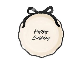 Puede incluir: Un plato de papel redondo de color crema con borde festoneado y contorno negro. El plato presenta un lazo decorativo negro sujeto al borde superior y el texto Happy Birthday impreso en letra cursiva negra en el centro de la superficie.