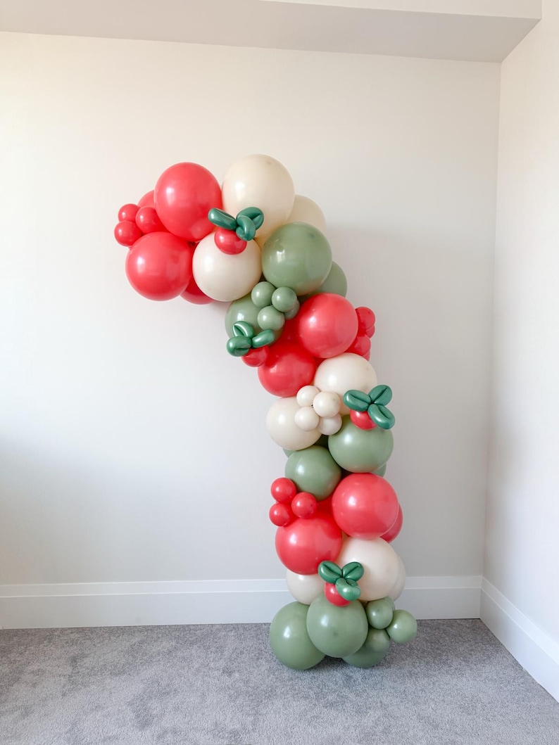 May include: A festive balloon arch in a curved shape, featuring red, cream, and green balloons. The balloons are arranged to resemble a holiday garland, with small green accents. The arch is set against a white wall and gray carpet.
