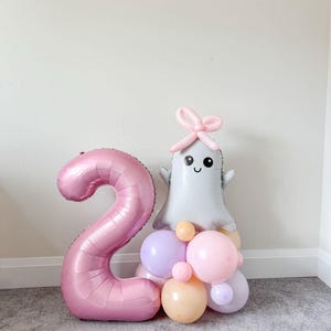 May include: A large pink number 2 foil balloon is positioned beside a ghost-shaped balloon with a bow. The ghost balloon is grey with a smiling face, and is surrounded by a cluster of pastel balloons in pink, lilac, and peach. A birthday party decoration.