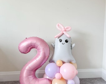 May include: A pink number 2 foil balloon stands next to a ghost-shaped balloon with a bow. The ghost balloon is gray with a smiling face and is surrounded by a cluster of pastel-colored balloons in pink, purple, and orange. A festive birthday decoration.