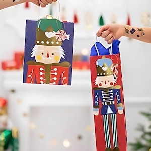Könnte beinhalten: Zwei rote und blaue Geschenktüten mit einem Nussknacker-Design. Die Taschen sind mit goldenen Akzenten verziert und haben einen Zuckerstock und eine Krone auf dem Kopf des Nussknackers.