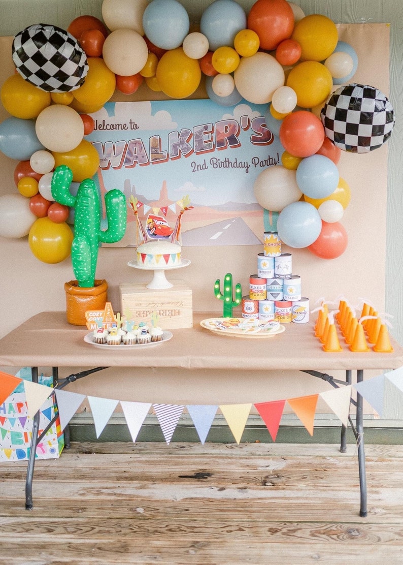 May include: A festive birthday party setup with a table decorated with balloons, a cake, cupcakes, and party favors. The backdrop reads "Welcome to Walker's 2nd Birthday Party!" with a road design. Includes a cactus and a banner.