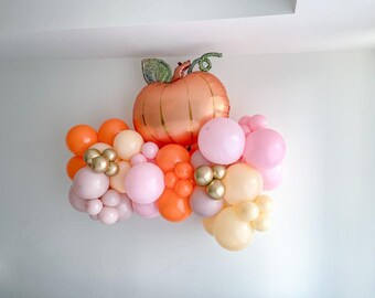 Könnte beinhalten: Ein großer orangefarbener Kürbisballon mit grünem Stiel und Blättern ist von einer Gruppe kleinerer Ballons in Rosatönen, Pfirsich und Gold umgeben.