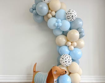 May include: A balloon arch made of blue, beige, and white balloons with paw print designs. A brown and beige dachshund balloon is in the foreground.