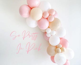 May include: A balloon garland featuring pink, white and beige balloons. The garland is shaped like an arch and is hanging on a white wall. The text "So Pretty In Pink" is written on the wall below the garland.