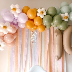 May include: A balloon arch made of pastel pink, lavender, and green balloons with white daisy-shaped balloons. The arch is decorated with orange and yellow balloons. There are two large brown balloons in the shape of the number 10. The balloons are hanging from a wall with a backdrop of pastel coloured streamers.
