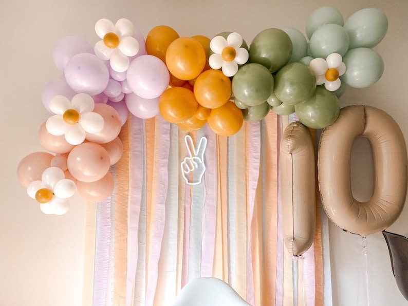 May include: A balloon arch made of pastel pink, lavender, and green balloons with white daisy-shaped balloons. The arch is decorated with orange and yellow balloons. There are two large brown balloons in the shape of the number 10. The balloons are hanging from a wall with a backdrop of pastel coloured streamers.