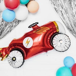 May include: A red race car shaped balloon with a white lightning bolt and black wheels. The balloon is hanging from a silver foil curtain with other balloons in the background.