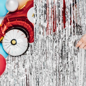 May include: Silver tinsel curtain with a hand pulling it open to reveal red, blue, and white balloons. The balloons are shaped like a car and a circle.