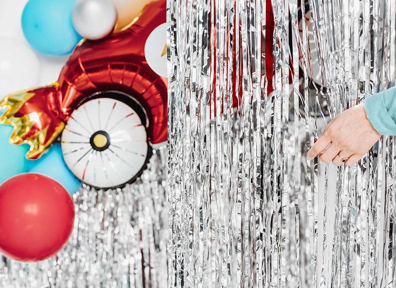 May include: Silver tinsel curtain with a hand pulling it open to reveal red, blue, and white balloons. The balloons are shaped like a car and a circle.