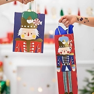 Könnte beinhalten: Zwei rote und blaue Geschenktüten mit einem Nussknacker-Motiv. Die Taschen sind mit goldenen Verzierungen und Zuckerstangen-Akzenten verziert.