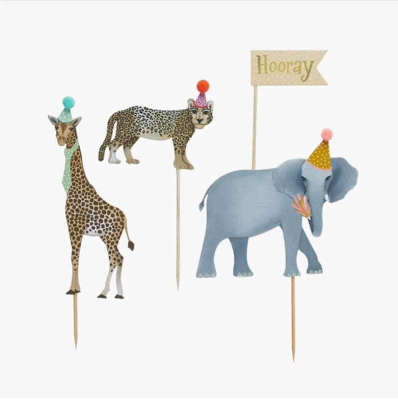 May include: A set of four cake toppers featuring a giraffe, a leopard, an elephant, and a banner that says "Hooray". Each animal is wearing a party hat and is made of paper.