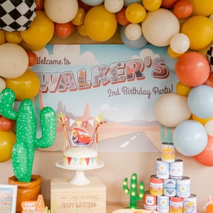 May include: A festive party scene with a "Walker's 2nd Birthday Party" backdrop, balloons in yellow, white, orange, and blue, and a cake with a Cars theme. Decorations include cactus inflatables and a stack of themed cans.