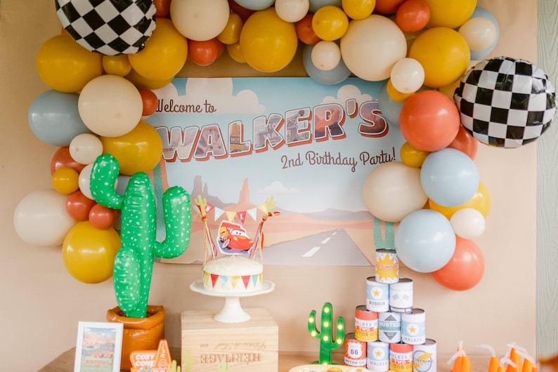 May include: A festive party scene with a "Walker's 2nd Birthday Party" backdrop, balloons in yellow, white, orange, and blue, and a cake with a Cars theme. Decorations include cactus inflatables and a stack of themed cans.