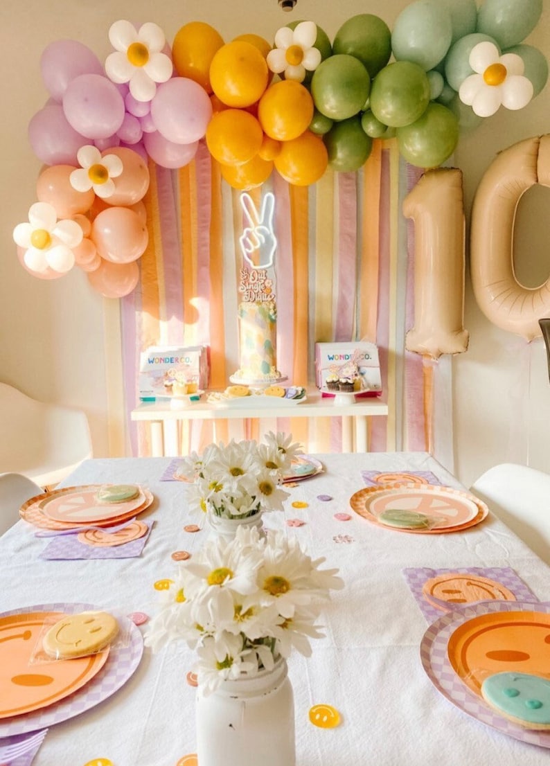 May include: A birthday party table setting with a white tablecloth, colourful plates featuring peace signs, and white daisies in mason jars. The backdrop features a rainbow of streamers and balloons with daisies. The cake is decorated with a peace sign and the words "Let's Get Funky 10 Years".