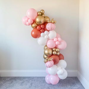 Puede incluir: Una guirnalda de globos vertical, con globos rosas, blancos, dorados y óxido. La guirnalda incluye globos decorativos con forma de calabaza. Los globos están dispuestos contra una pared blanca.