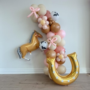 May include: A balloon arch featuring a brown horse, a gold horseshoe, and pink, brown, and white balloons. The balloons are decorated with cow print and flowers.