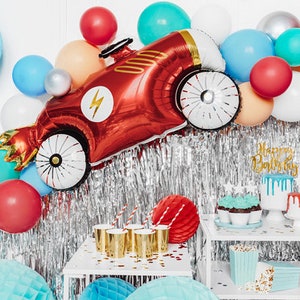 May include: A red race car shaped balloon with a lightning bolt design, surrounded by colorful balloons and a silver tinsel backdrop. There are two white shelves with cupcakes, a cake, and popcorn containers. The top shelf has a gold banner that says "Happy Birthday".