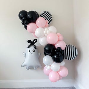 May include: A festive balloon garland in black, white, pink, and striped patterns. A silver ghost-shaped balloon with a black bow and smiling face is also present. The balloons are arranged on a white wall.