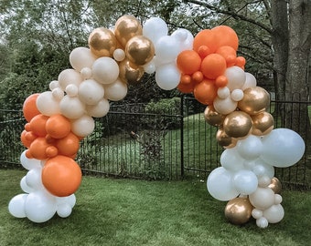 Könnte beinhalten: Ein Ballonbogen aus weißen, beigen, goldenen und orangen Ballons. Der Bogen befindet sich vor einem schwarzen Metallzaun und grünem Gras.