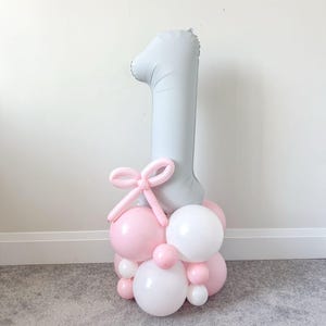 May include: A festive balloon arrangement for a first birthday celebration. It features a large, light gray number "1" balloon, a pink bow, and a cluster of pink and white balloons of various sizes. The arrangement is set on a gray carpet.