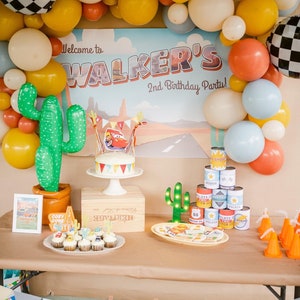 May include: A festive birthday party scene with balloons, a cake, and decorations. The backdrop reads "Welcome to WALKER'S 2nd Birthday Party!" A cake with a Cars theme sits on a white stand. Other decorations include cactus balloons and cupcakes.