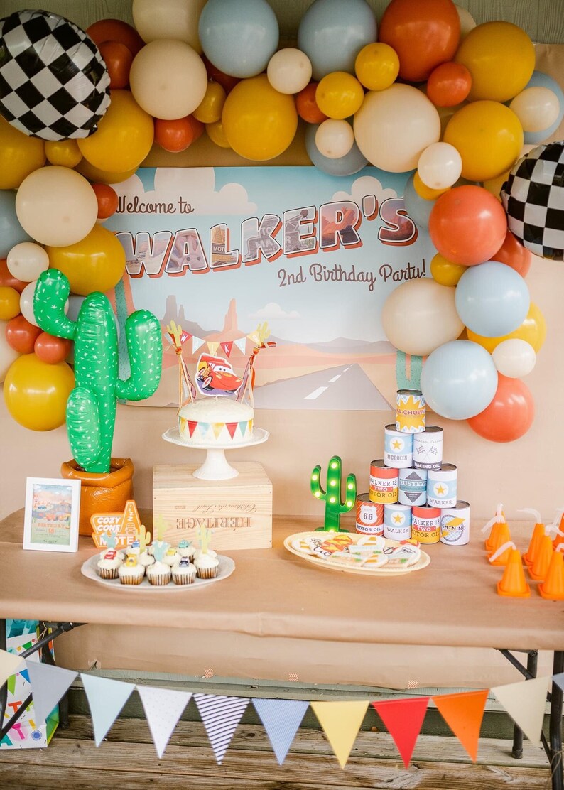 May include: A festive birthday party scene with balloons, a cake, and decorations. The backdrop reads "Welcome to WALKER'S 2nd Birthday Party!" A cake with a Cars theme sits on a white stand. Other decorations include cactus balloons and cupcakes.