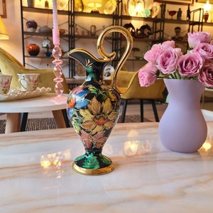 Könnte beinhalten: Eine schwarze und goldene Keramikkanne mit Blumenmuster. Die Kanne hat eine breite Öffnung und einen Henkel. Sie steht auf einem Tisch mit einer Vase mit rosa Blumen.