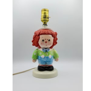 May include: A ceramic lamp shaped like a rag doll with red hair, a green and blue dress, and a white base. The lamp has a gold lightbulb.