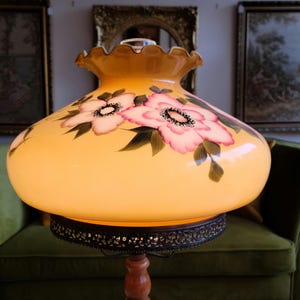 May include: A vintage table lamp with a yellow glass shade featuring hand-painted pink and white flowers. The shade has a scalloped edge and sits on a wooden base.