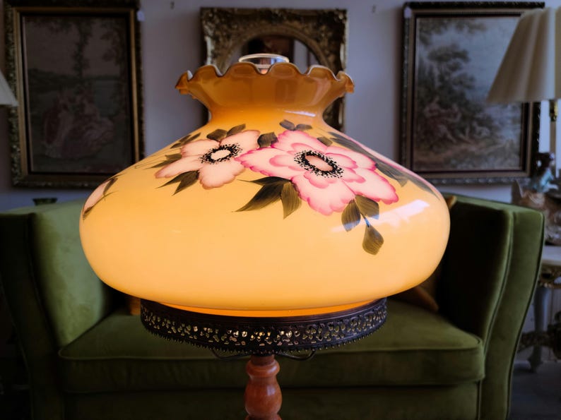 May include: A vintage table lamp with a yellow glass shade featuring hand-painted pink and white flowers. The shade has a scalloped edge and sits on a wooden base.
