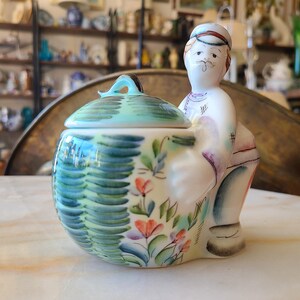 May include: A ceramic sugar bowl with a lid, featuring a whimsical painted design of a person with a hat and a floral pattern. The bowl is painted in shades of green, blue, pink, and white.