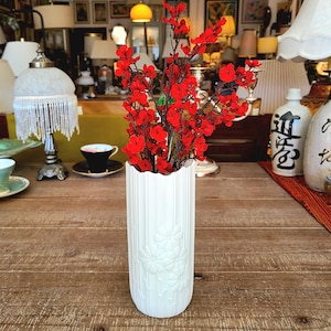May include: White ceramic vase with an embossed floral design, holding a bouquet of red artificial flowers.