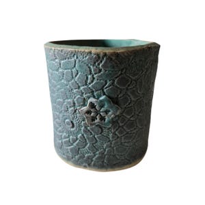 Op de afbeelding: Een turquoise keramische beker met een getextureerd, patroon oppervlak. Een sneeuwvlokvormig ornament is aan de voorkant bevestigd. De beker heeft een enigszins onregelmatige vorm en een donkere rand.