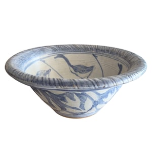 May include: A ceramic bowl featuring a wide, textured rim with blue and white brushstroke patterns. The interior and exterior surfaces are decorated with hand-painted blue illustrations of long-necked birds and foliage against a cream base.