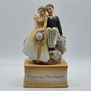 May include: A porcelain figurine depicting two figures sitting on a rock, mounted on a rectangular base with the text Norman Rockwell. The figure on the left wears a yellow dress and holds a straw hat, while the figure on the right wears a dark jacket.