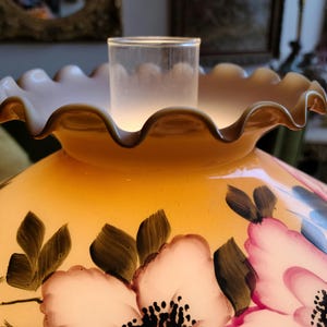 May include: A yellow glass lampshade with a ruffled edge and a floral design. The flowers are painted in shades of pink, white, and green.