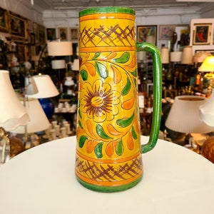 May include: A yellow ceramic jug with a green handle and floral designs in orange, green, and brown. The jug has a lattice pattern around the top and bottom.