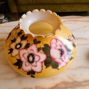 May include: A yellow glass lampshade with a white ruffled top and painted pink and white flowers with green leaves.