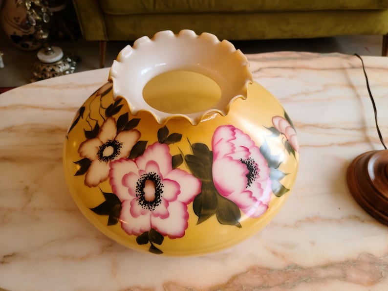 May include: A yellow glass lampshade with a white ruffled top and painted pink and white flowers with green leaves.